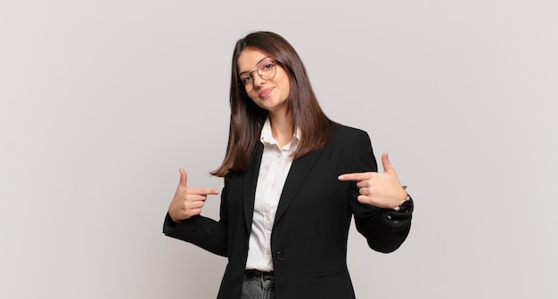 Mujer de negocios joven que parece orgullosa, arrogante, feliz, sorprendida y satisfecha, apuntando a sí misma, sintiéndose como una ganadora