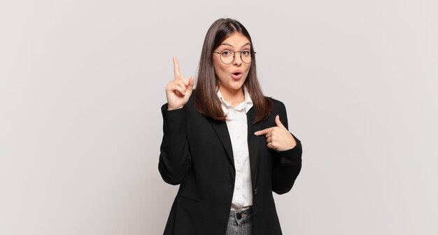 Mujer de negocios joven que se siente orgullosa y sorprendida, apuntando a sí misma con confianza, sintiéndose como el número uno exitoso