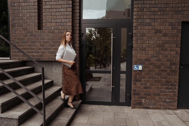 Mujer de negocios joven que sostiene la computadora portátil del fondo del edificio de oficinas moderno