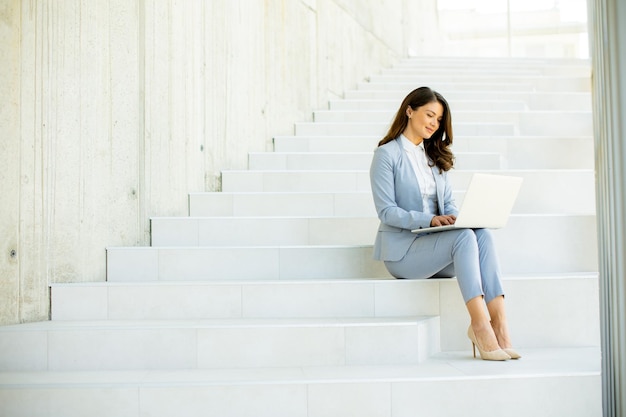 Mujer de negocios joven sentada en las escaleras en el pasillo de la oficina y trabajando en la computadora portátil