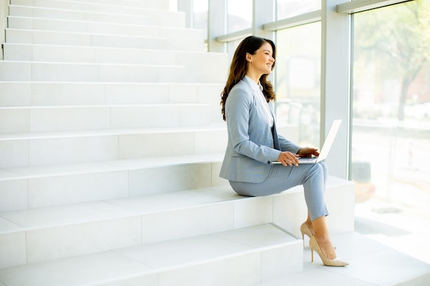 Mujer de negocios joven sentada en las escaleras en el pasillo de la oficina y trabajando en la computadora portátil