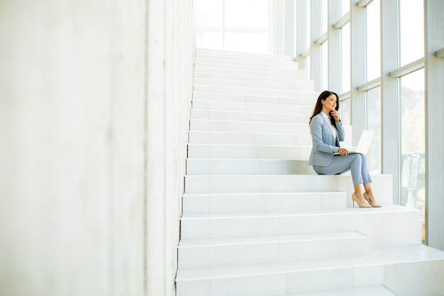 Mujer de negocios joven sentada en las escaleras en el pasillo de la oficina y trabajando en la computadora portátil