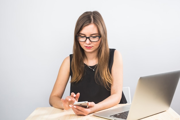 Mujer de negocios joven sentada en la oficina en la mesa y con smartphone. En el escritorio hay una computadora portátil. Mujer analizando datos. Aprendizaje de estudiantes en línea.