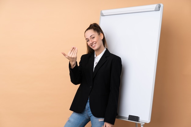 Mujer de negocios joven sobre la pared aislada que da una presentación en el tablero blanco e invita a venir con la mano