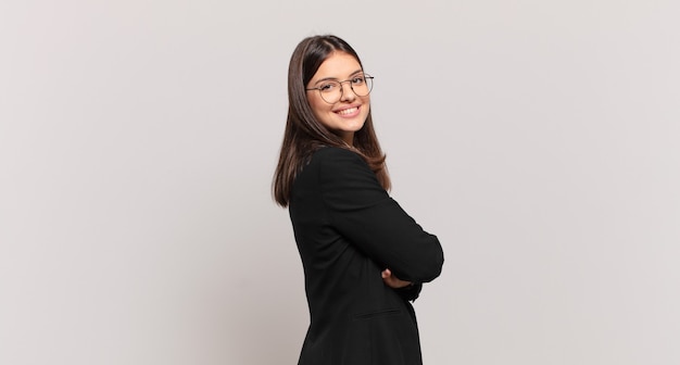 Mujer de negocios joven sonriendo alegremente, sintiéndose feliz, satisfecha y relajada, con los brazos cruzados y mirando hacia un lado