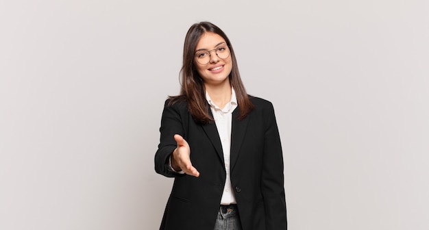 Mujer de negocios joven sonriendo, luciendo feliz, confiada y amigable, ofreciendo un apretón de manos para cerrar un trato, cooperando