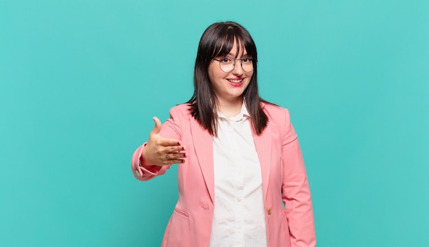 Mujer de negocios joven sonriendo, luciendo feliz, confiada y amigable, ofreciendo un apretón de manos para cerrar un trato, cooperando