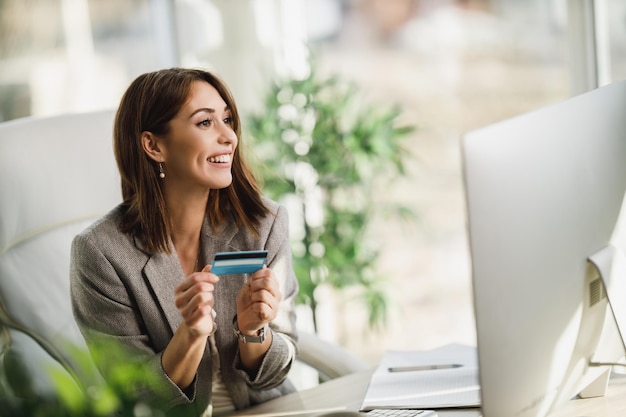 Una mujer de negocios joven sonriente que usa una computadora y una tarjeta de crédito en la oficina.