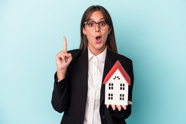 Mujer de negocios joven sosteniendo una casa de juguete aislada sobre fondo azul con una gran idea, concepto de creatividad.