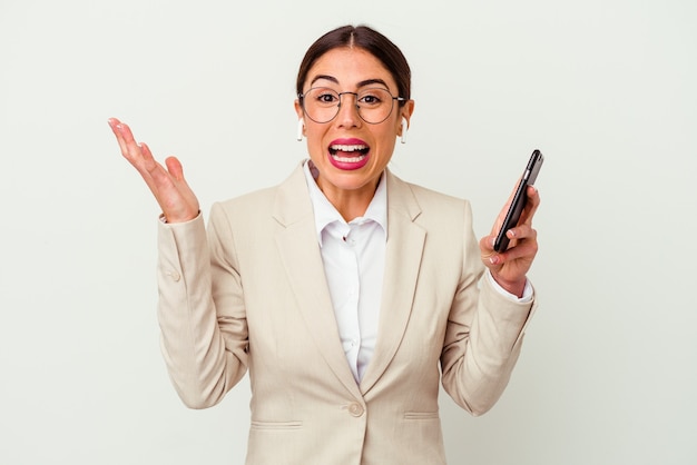 Mujer de negocios joven sosteniendo un teléfono móvil aislado en blanco recibiendo una agradable sorpresa, emocionada y levantando las manos.