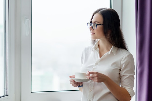 Mujer de negocios joven con una taza de café cerca de la ventana en la oficina, ciudad en la ventana para la temporada de otoño invierno. Coffee break, profesora positiva, psicóloga, consultora, secretaria