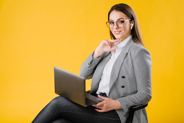 Mujer de negocios joven en traje con gafas sentada y usando una computadora portátil sobre fondo amarillo