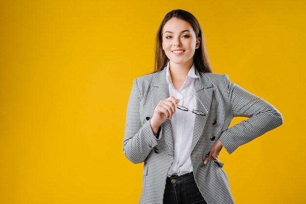 Mujer de negocios joven en traje sosteniendo anteojos y mirando a la cámara sobre fondo amarillo