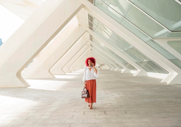 Mujer de negocios latina de moda y sonriente con cabello afro rojo hablando en su teléfono inteligente mientras camina bajo una arquitectura moderna en forma de triángulo