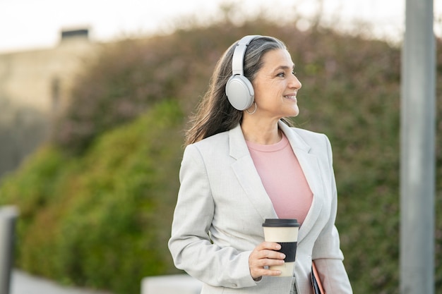 Mujer de negocios madura con café y auriculares escucha música en la ciudad