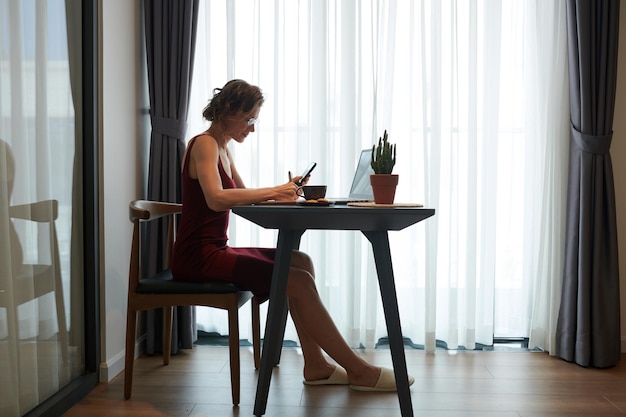 Mujer de negocios madura y elegante con anteojos sentada en el escritorio en la sala de estar revisando las notificaciones en pho