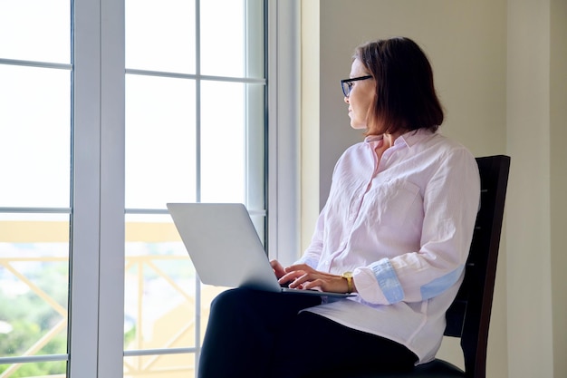 Mujer de negocios madura con gafas con portátil en las manos cerca de la ventana