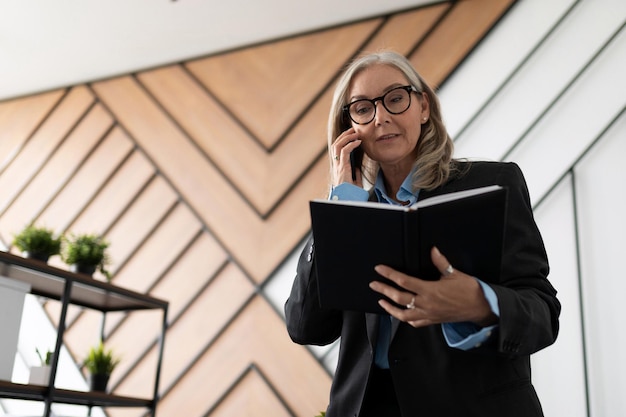 Mujer de negocios madura con gafas y ropa de negocios habla por teléfono móvil con un bloc de notas en ella