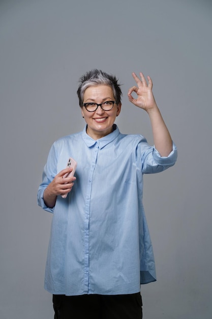 Mujer de negocios madura de pelo gris gesticulando bien con el teléfono inteligente en la mano mirando a la cámara con camisa azul Mujer bonita con camisa azul aislada en blanco Imagen tonificada