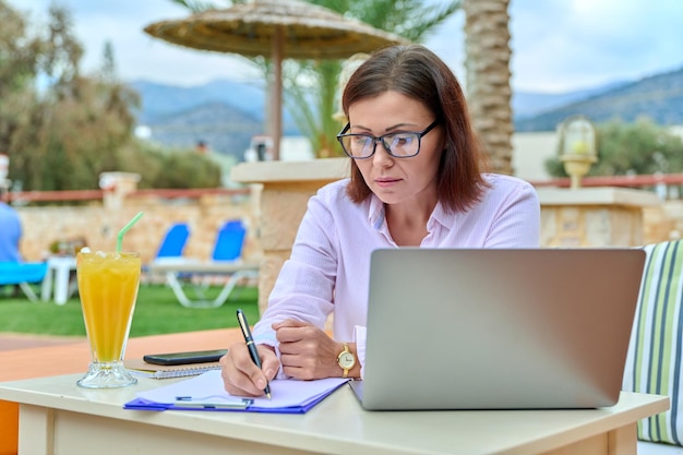 Mujer de negocios madura que trabaja con una laptop sentada al aire libre cerca de la piscina