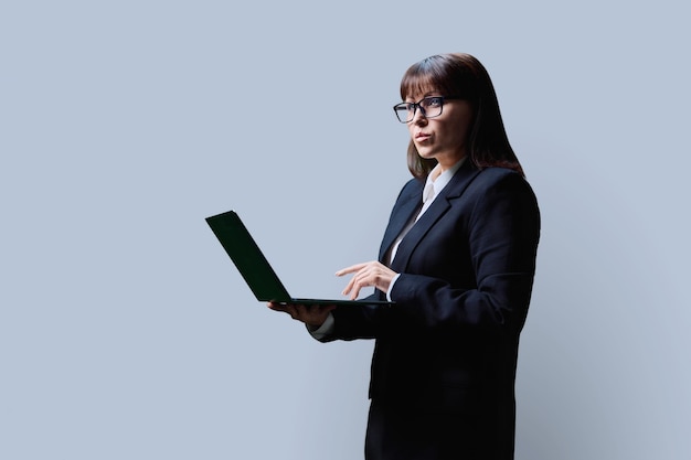 Una mujer de negocios madura y seria usando una computadora portátil sobre un fondo gris.