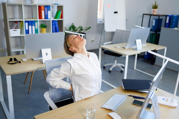 Foto mujer de negocios madura se sienta en su lugar de trabajo en la oficina y se aferra a su espalda baja