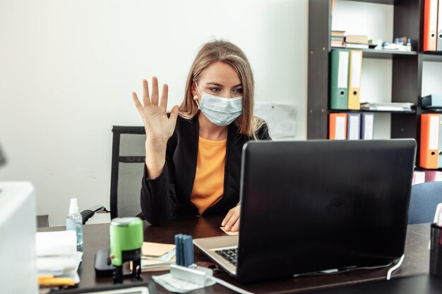 La mujer de negocios con una máscara médica se comunica por videollamada en la oficina en el lugar de trabajo