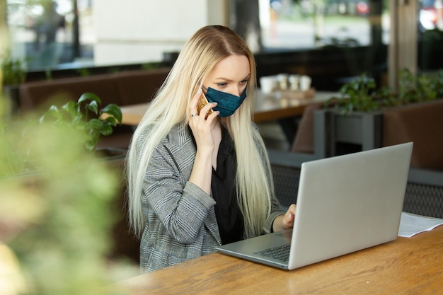 Mujer de negocios con máscara médica usa una computadora portátil y habla por teléfono mientras está sentada en la mesa en un café al aire libre Trabajo remoto Pandemia de Covid19