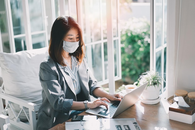 Mujer de negocios con máscara en ropa informal inteligente que trabaja en una computadora portátil mientras está sentada en una oficina creativa o una mujer de café que trabaja con una computadora portátil en la mesa de madera en la cafetería