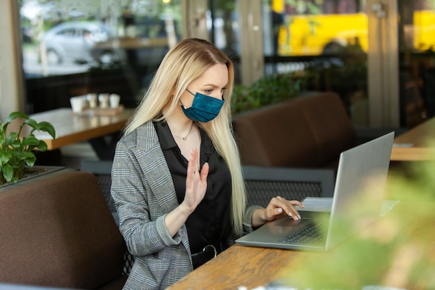 Mujer de negocios con mascarilla médica saludando con la mano en la pantalla del portátil dando la bienvenida a los interlocutores por videoconferencia mientras se sienta a la mesa en un café al aire libre Teletrabajo pandemia de coronavirus