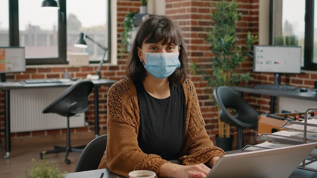 Mujer de negocios con mascarilla y portátil en la oficina de inicio. Retrato de un empleado de la organización que trabaja en la planificación de proyectos y la estrategia de marketing con una computadora durante una pandemia.