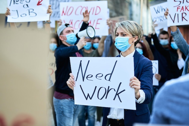 Mujer de negocios con mascarilla protectora que lleva una pancarta con la inscripción Need Work mientras protesta con la multitud en las calles