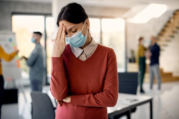 Mujer de negocios con mascarilla sosteniendo su cabeza con dolor mientras está de pie en la oficina