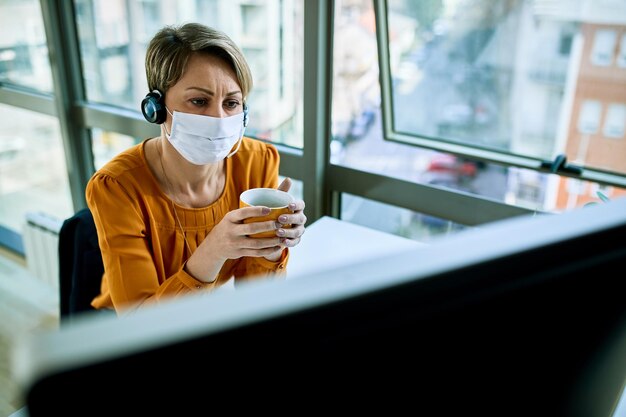 Mujer de negocios con mascarilla sosteniendo una taza de café mientras lee un correo electrónico problemático en una PC de escritorio en la oficina