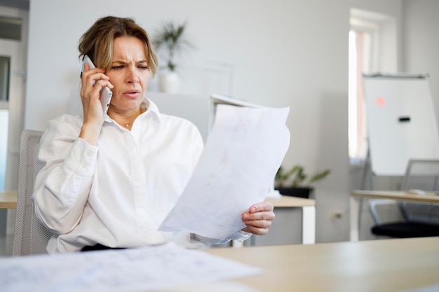 Mujer de negocios de mediana edad con cara seria hablando en un teléfono inteligente mientras mira documentos