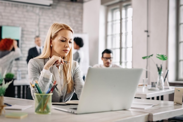 Mujer de negocios de mediana edad que usa una computadora portátil y lee un correo electrónico en el trabajo Hay personas en el fondo