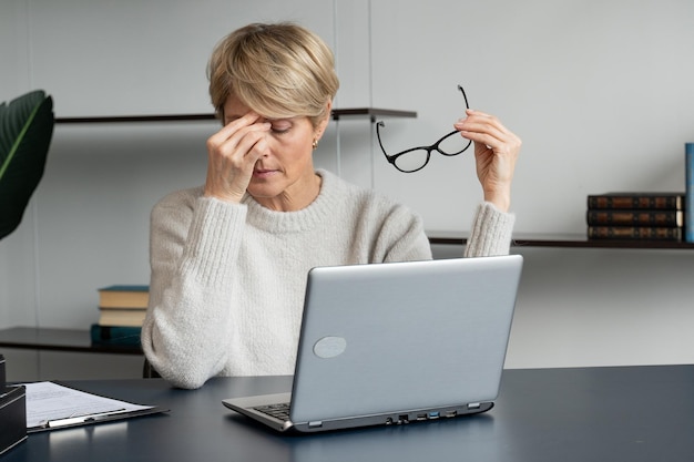 Una mujer de negocios de mediana edad sentada en su escritorio se quita las gafas por el dolor en sus ojos y