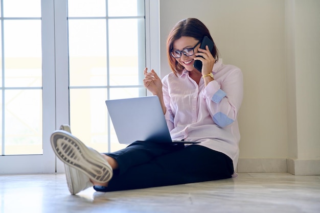 Mujer de negocios de mediana edad sentada en el suelo con una laptop hablando por un teléfono móvil