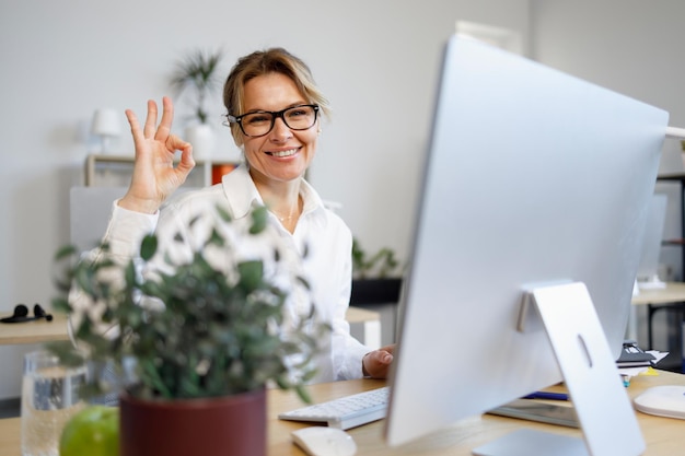 Una mujer de negocios de mediana edad se sienta frente a una computadora en la oficina y muestra un gesto correcto con los dedos