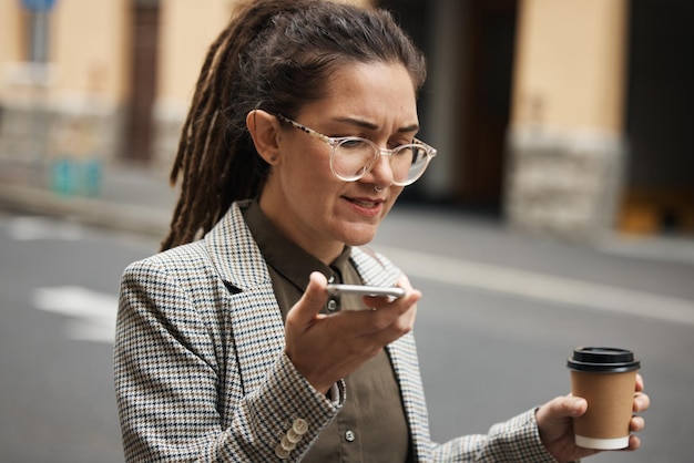 Mujer de negocios en un mensaje de voz en una ciudad para viajar o viajar por la mañana con café hablando para contactar Hipster en línea y empleado caminando hablando o trabajando en red con teléfono celular en internet