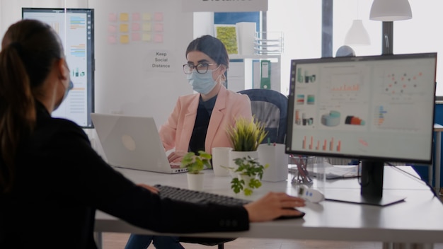 Mujer de negocios mirando gráficos financieros en la pantalla de la computadora mientras habla con un compañero de trabajo sentado en la oficina de la empresa. Compañeros con mascarillas que mantienen el distanciamiento social para prevenir la enfermedad covid19