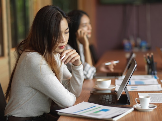 mujer de negocios, mirar, informe financiero, en, el, escritorio, con, computadora portátil, material de oficina, y, compañero de trabajo