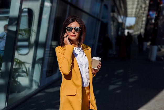 Mujer de negocios moderna en gafas de sol y traje amarillo se encuentra cerca del centro de la oficina con una taza de café y habla en el teléfono inteligente. Concepto de estilo de vida y éxito empresarial.