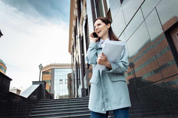 Mujer de negocios moderna hablando por teléfono mientras sostiene una carpeta con documentos en las escaleras