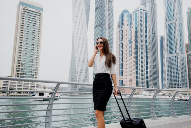 Mujer de negocios moderna joven ocupada en traje tirando de una maleta en un marine de Dubai. Caminando hacia su oficina y hablando por teléfono.