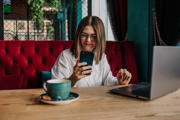 Una mujer de negocios morena está sentada en una mesa en un café con una computadora portátil y mirando su teléfono