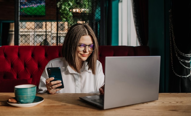 Una mujer de negocios morena con gafas está sentada en una mesa en un café con un teléfono y trabajando en una computadora portátil