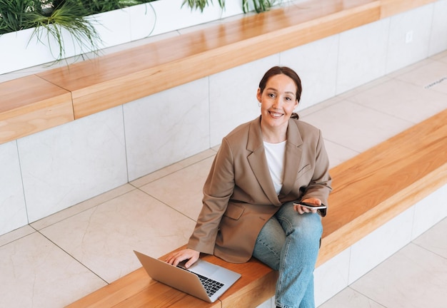 Mujer de negocios morena sonriente adulta de cuarenta años con cabello largo en elegante traje beige y jeans trabajando en una laptop en un lugar público