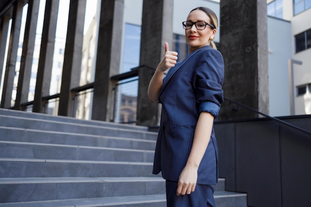 Mujer de negocios mostrando los pulgares hacia arriba mientras está de pie en las escaleras cerca del edificio de oficinas