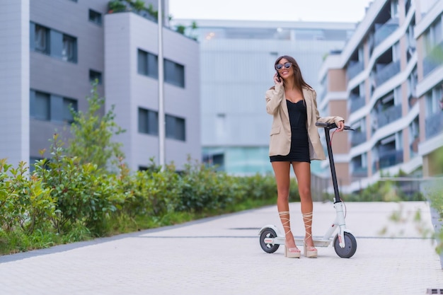 Una mujer de negocios moviéndose con un scooter eléctrico por la ciudad gafas de sol sonriendo hablando por teléfono